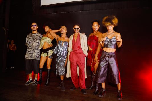 Foto de um grupo de seis pessoas negras, todas de pé. Algumas olham para a câmera e outras não. Três delas estão de roupas pretas, com alguns detalhes em branco, que remetem à arte do grafite de rua. Uma das pessoas usa peças com as cores preto, verde e branco. As outras duas pessoas estão de vermelho. 