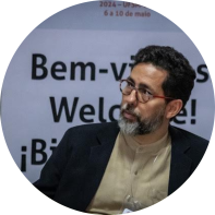 Foto de homem pardo com olhos escuros. Ele tem cabelos cacheados, curtos e escuros e barba e bigode grisalhos. Ele usa óculos com hastes vermelhas, camisa bege e blazer preto. No fundo, um painel com a inscrição “Bem-vindos” em diferentes idiomas.