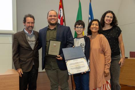 Foto em plano americano de cinco pessoas lado a lado, posando de pé e sorrindo. Um homem branco, de cabelos curtos e lisos e barba, ligeiramente grisalha, vestindo um blazer e calça marrom, camisa azul e blusa de lã cinza; um homem branco e calvo de olhos castanhos, cabelos curtos, lisos e castanhos e barba. Ele veste uma camisa e calça verde oliva e um paletó azul. Em uma das mãos, segura uma placa de premiação; um garoto branco, de cabelos curtos e pretos, usando óculos de armação escura. Veste uma camiseta manga longa preta e branca e segura uma pasta com um certificado; uma mulher branca, de cabelos castanhos, médios e lisos, vestindo um vestido longo estampado, em laranja e branco, e um casaco de lã laranja claro; e uma mulher branca, de cabelos longos, castanhos e ondulados, usando óculos de armação prata. Ela veste uma blusa preta e calça jeans cinza. Ao fundo, uma parede branca, móveis de madeira, e três bandeiras hasteadas.