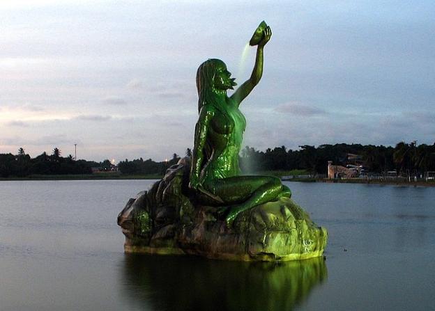 Foto de uma estátua de uma mulher nua de cabelos longos se banhando sentada em uma pedra. Em uma mão ela carrega uma lança e, na outra, que está erguida sobre a cabeça, um recipiente com água que ela derrama sobre seu corpo, como um chafariz. A estátua é iluminada de verde. Ao fundo, a água do lago, árvores em sua margem e o céu com nuvens.