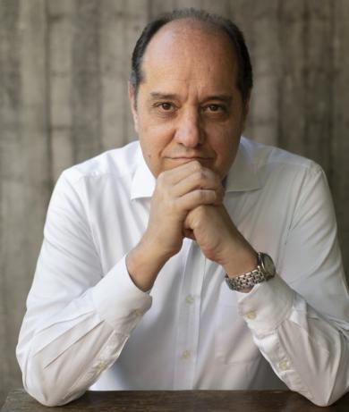 Foto de um homem branco, calvo, de cabelos escuros e curtos. Ele veste camisa social branca e relógio de pulso prateado. Sorri ligeiramente, com as mãos unidas sob o queixo e os cotovelos apoiados sobre uma mesa. O fundo é cinza e desfocado.