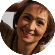 Descrição da imagem: Foto de mulher sorrindo e com a mão na cabeça. Ela veste blusa marrom, tem cabelos castanhos, lisos e curtos e olhos castanhos. Usa batom vermelho rosado. O fundo é branco.