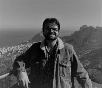 Pedro Guilherme é um jovem branco de cabelo e barba castanhos. Usa um óculos de haste preta. Na foto, usa uma jaqueta jeans e u