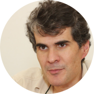 Foto de homem branco com olhos castanhos e cabelos curtos, ondulados e escuros com alguns fios grisalhos. Ele veste camisa bege e o fundo da imagem também tem a cor bege.