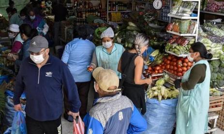 Foto de pessoas em um mercado durante a pandemia. Grande parte delas segura sacolas plásticas e todas utilizam máscaras. É possível observar alguns produtos à venda, como legumes e vegetais.