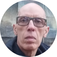 Foto de homem branco com expressão séria. Ele é careca, tem olhos castanhos e usa óculos de armação quadrada e escura. Ele usa uma blusa azul escura e o fundo da imagem é cinza azulado. 