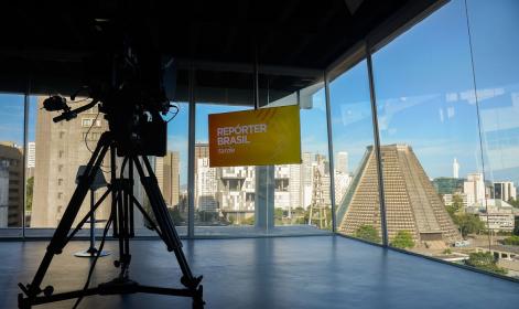 Foto de um estúdio cercado por janelas de vidro e localizado em um andar alto, com vista para a região central do Rio de Janeiro. Há um tripé com uma câmera no canto esquerdo do estúdio, que aponta na direção de um monitor de TV que exibe os dizeres &quot;Repórter Brasil&quot;. 