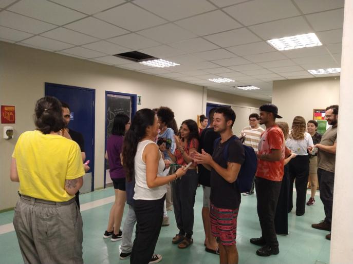 Na fotografia, cerca de quinze estudantes de diversos gêneros e cores estão em uma sala de piso verde e teto branco. Eles conversam em duplas ou pequenos grupos. 