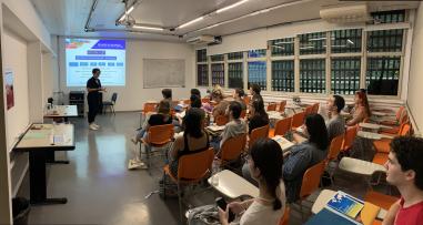 Foto colorida em ambiente interno, de uma sala de reunião com estudantes estrangeiros e a secretária da CRInt, Thaise Desirre, os recepcionando 