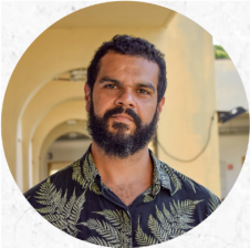 Foto de homem negro olhando para a câmera. Ele tem cabelos curtos, pretos, barba e bigode pretos. Veste uma camisa preta com estampa de folhas verdes. Atrás há um corredor com paredes e arcos amarelo-claro.