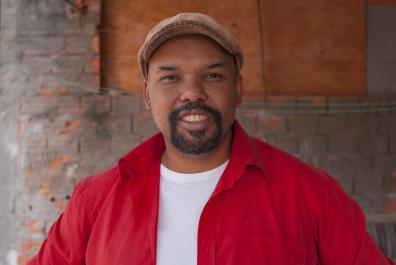 Foto de um homem de pele negra e barba escura, sorrindo. Ele veste uma boina bege e uma camisa vermelha por cima de uma camiseta branca. Ao fundo, atrás do homem, uma parede de tijolos laranjas cobertos por cimento. Uma parte da parede é substituída por um pedaço retangular de madeira.