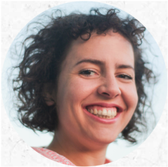 Foto de uma mulher branca olhando para a câmera e sorrindo. Ela tem cabelos pretos, cacheados na altura da nuca. Atrás dela, um céu azul.
