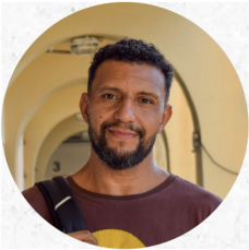 Foto de um homem negro esboçando um sorriso. Ele tem cabelos curtos pretos, barba e bigode pretos. Veste uma camisa roxa. Atrás dele há a parte superior de um corredor com paredes e arcos amarelo-claro.