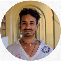 Foto de um homem negro sorrindo. Ele tem cabelos cacheados, curtos e pretos, bigode preto e sinais de barba. Veste uma camisa branca, utiliza um brinco na orelha direita, corrente dourada e colar azul com pingente de búzio. Atrás dele há um corredor com paredes e arcos amarelo-claro.