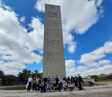 Foto colorida em ambiente externo, de estudantes estrangeiros, junto das Comissões Internacionais citadas na notícia, em frente à torre do relógio da Praça do Relógio na USP