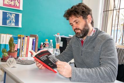 Foto de Thiago Mio Salla lendo o livro O Antimodernista. A foto é tirada do lado esquerdo, mas é possível ver os dois olhos do professor. Um homem branco de barba e cabelos curtos castanhos ondulados. Do lado direito há uma fileira de livros posicionados sobre a mesa, uma parede azul com alguns desenhos colados e uma janela com grade ao fundo