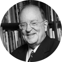 Foto em preto e branco de um homem branco sorrindo. Ele é calvo, usa óculos com hastes finas e armação transparente. Ele veste paletó preto, camisa branca e gravata. Ao fundo, uma estante de livros.