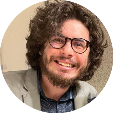 Homem branco, usando óculos. Tem barba e cabelos grandes. Está sorrindo, de frente. Veste uma camisa azul com gola, por cima, um terno bege. Ao fundo, uma parede bege borrada.