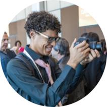 Descrição: Foto de um rapaz negro de cabelos curtos, enrolados e castanhos. Ele está de perfil, usa óculos, sorri, segura uma câmera fotográfica e veste camisa azul escura de mangas longas e gravata cor de rosa. Ao fundo, algumas pessoas aparecem desfocadas.