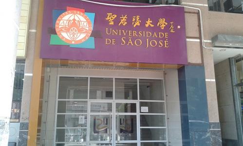 Foto de uma porta de vidro, entremeada de ferragens brancas. Ela está fechada. Acima, encontra-se uma placa roxa, com o nome do local (Universidade de São José) em português e em ideogramas chineses. Ao lado, na própria placa, está o logo da instituição.