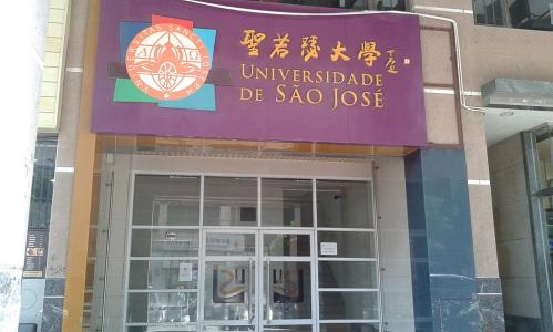 Foto de uma porta de vidro, entremeada de ferragens brancas. Ela está fechada. Acima, encontra-se uma placa roxa, com o nome do local (Universidade de São José) em português e em ideogramas chineses. Ao lado, na própria placa, está o logo da instituição. 