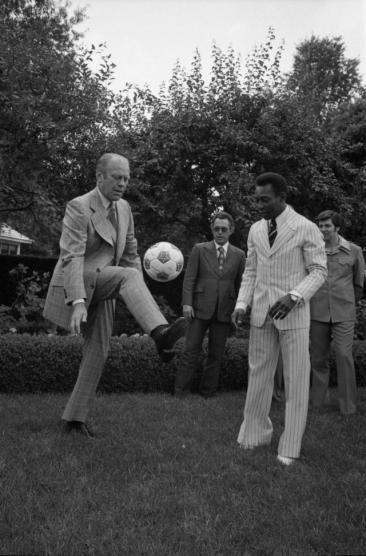 Foto em preto e branco de Pelé e Gerald Ford. O primeiro, negro de cabelos crespos e curtos, veste um terno branco risca de giz e uma gravata preta, juntamente com um sapato branco. Já o segundo, um homem branco e idoso, veste um terno cinza xadrez com um colete da mesma estampa, além de sapatos sociais pretos. Ele faz embaixadinhas com uma bola. Atrás deles, dois homens observam a cena. Os quatro homens encontram-se em um gramado, o qual parece fazer parte de um jardim. 