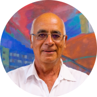 Foto de um homem branco. Ele é calvo, tem cabelos brancos e curtos nas laterais, tem olhos castanhos, veste camisa branca e usa óculos. Ao fundo uma parede colorida em tons de vermelho, azul, laranja, verde e branco.