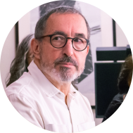 Foto de homem branco com olhos castanhos, cabelos curtos e grisalhos e barba grisalha. Ele usa óculos de armação redonda e preta, camisa branca e olha para o lado com expressão neutra. Ao fundo, pessoas e um quadro em uma parede branca. 