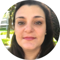 Foto de mulher branca com olhos castanhos e cabelos lisos e castanhos abaixo dos ombros. Ao fundo, arbustos e árvores.