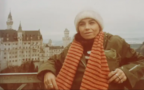 Foto de uma mulher branca, vestindo cachecol laranja listrado, um casaco verde pesado e gorro branco. Ela está apoiada sobre uma grade. Atrás dela, encontra-se uma grande edificação com torres, que lembra um castelo.