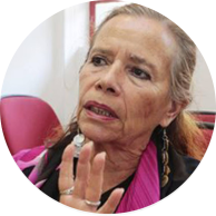 Foto de uma mulher parda de olhos castanhos, cabelos longos, ondulados, castanhos e grisalhos. Ela olha para um dos lados da imagem, falando e gesticulando com a mão aberta, usa brincos longos, anéis, blusa preta e lenço cor de rosa. Ao fundo, há objetos vermelhos e pretos e uma parede branca.