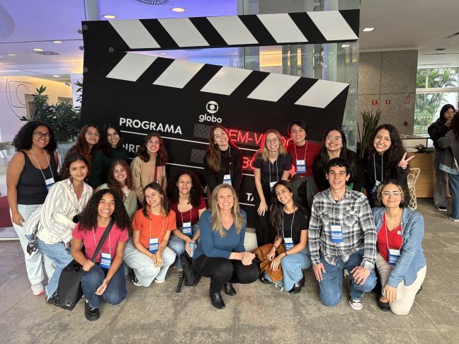 Foto de um grupo de cerca de 20 pessoas, em sua maioria mulheres jovens e brancas, todos com um crachá no pescoço. Alguns estão de pé e outros agachados na frente de uma claquete gigante com o logo da Rede Globo em um espaço com piso de cimento e divisórias em vidro ao fundo.