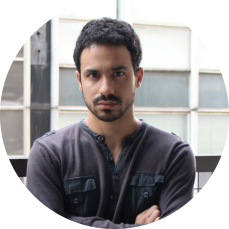 Homem branco, de cabelo preto curto e barba. Tem olhos castanhos e olha para a frente. Veste uma camisa de botões e manga longa azul escura. Está de braços cruzados, parado em uma sacada. Ao fundo, as janelas de um prédio.