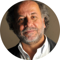 Foto de homem branco esboçando sorriso. Ele tem  olhos castanhos, cabelos curtos, cacheados e grisalhos, barba, bigode grisalhos e usa camisa branca e casaco preto. O fundo é marrom.