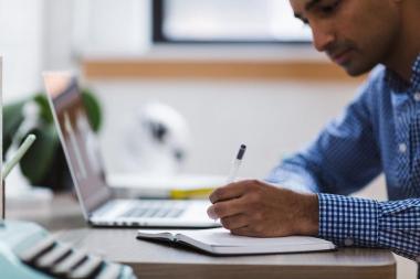 Um homem branco, de camisa xadrez azul e branca, escreve com uma caneta em um caderno. O caderno está sobre uma mesa, onde também se encontra um laptop aberto.