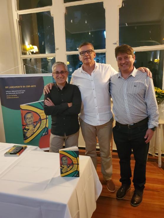 Foto de três homens brancos, abraçados, em pé, sorrindo. Ao fundo uma parede branca com janelas e o cartaz de lançamento do livro. Da esquerda para a direita: Marcelo Cardoso, homem branco com cabelos grisalhos. Veste camisa preta, calça bege, está de braços cruzados e usa óculos; Miguel Ângelo da Luz, homem branco de cabelos grisalhos, usa óculos e veste camisa branca com calça bege e sapatos marrons; Luciano Maluly, homem branco, cabelos castanhos, veste camisa listrada em preto e branco, calça preta e sapatos pretos. À frente, há uma mesa com um pano branco e o livro em cima.