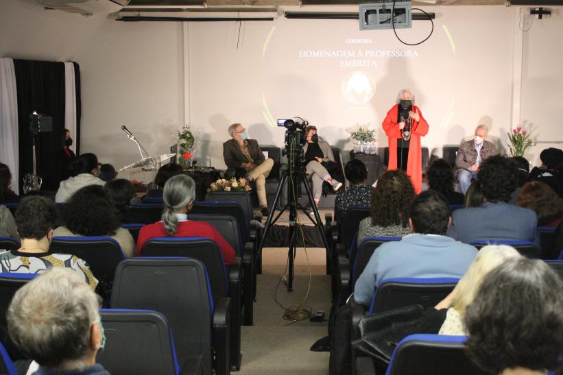 Foto do auditório do Departamento de Jornalismo e Editoração da ECA. Ao fundo, uma tela de projeção com uma foto da professora Ana Mae Barbosa. A professora, usando vestes pretas e um casaco vermelho, fala ao microfone. Atrás dela encontram-se sentados a diretora e o vice-diretor da ECA. O primeiro plano da imagem mostra pessoas sentadas em cadeiras azuis e de costas para a câmera enquanto assistem a cerimônia. Todos usam máscaras no rosto. 