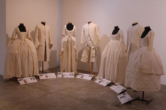Foto de roupas brancas, masculinas - calças e casacos - e femininas - vestidos -, de estilo antigo, dispostas em semicírculo em manequins pretos sem cabeça. Aos pés de cada manequim, um painel impresso com um descritivo do traje