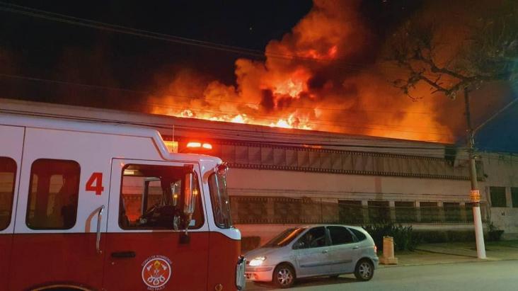 No canto esquerdo, parte frontal de um caminhão do corpo de bombeiros. Ao fundo, o galpão da Cinemateca Brasileira, um prédio de dois andares, é consumido pelo fogo