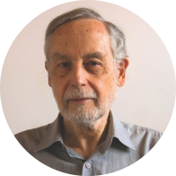 Foto de homem branco idoso. Ele tem olhos castanhos, cabelos lisos, curtos e grisalhos e barba grisalha. Ele veste camisa cinza e o fundo da imagem é de cor clara.