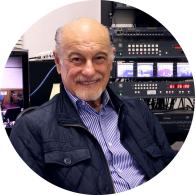 Foto de um homem branco sorrindo para a câmera. Ele é calvo e tem barba grisalha, veste uma camisa azul listrada e um casaco azul escuro. No fundo há equipamentos de gravação. 