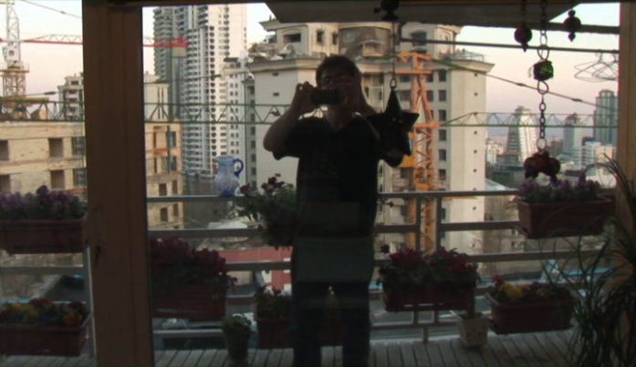 Foto de um homem segurando um celular na horizontal na altura de seu rosto. Ele está em pé em uma varanda, que está cercada por grades, onde estão pendurados diversos vasos com flores. Ao fundo, estão vários prédios, alguns em obras. O homem tem cabelos curtos e veste calça e camiseta, mas não é possível ver detalhes porque ele está contra a luz, o que deixa sua imagem escura.