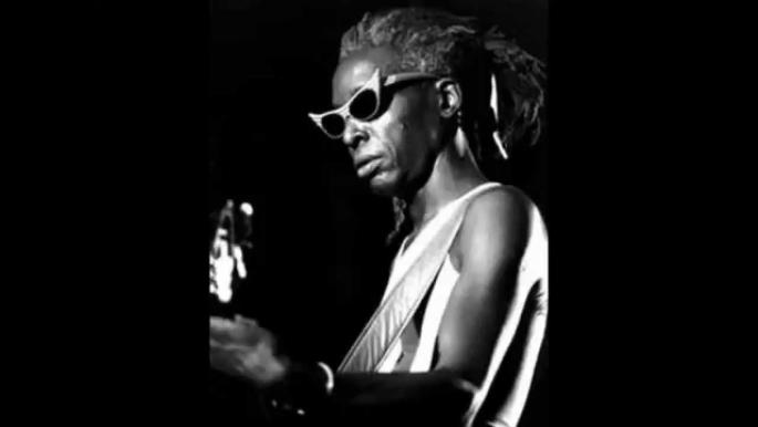 Foto em preto e branco de pessoa com cabelo preso em dreadlocks e óculos escuros angulosos tocando uma guitarra. Veste regata clara e está iluminada por um foco de luz, contrastando com o fundo escuro.