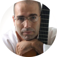 Foto de um homem branco que sorri ligeiramente e abraça um violão, tendo o braço do instrumento na vertical, ao lado do rosto. Ele tem olhos castanhos, é calvo e tem cabelos raspados bem curtos nas laterais. Veste camisa branca e usa óculos de armação fina. O fundo é branco.