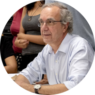 Foto de semi-perfil de um homem branco de cabelos brancos, ondulados e curtos e de olhos castanhos. Ele usa óculos sem aros e de hastes pretas e relógio de pulso,  Veste camisa azul clara, sorri ligeiramente e tem os antebraços apoiados sobre uma mesa. Ao fundo, há pessoas em pé.