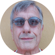 Foto de um homem branco de cabelos curtos, lisos  e grisalhos. Ele usa óculos de armação oval e lentes um pouco escuras,  camiseta vermelha e colar de contas brancas. Atrás dele há uma parede amarela.