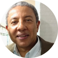 Foto de homem negro sorrindo. Ele tem cabelos curtos e grisalhos, olhos verdes e veste camisa branca e paletó marrom. Atrás dele, uma parede branca.
