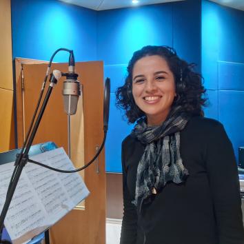 Foto de mulher branca sorrindo em estúdio de gravação. Seus cabelos são escuros, de tamanho médio e ondulados e ela veste uma camisa preta de mangas longas com cachecol xadrez verde. À sua esquerda há um microfone e uma partitura musical. O fundo é azul com uma porta de madeira clara.