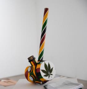 Foto de uma obra de arte composta por um tubo e outros acessórios de pvc formando uma estrutura que lembra um cachimbo gigante. O tubo é pintado com listras nas cores vermelho, amarelo e verde, e o ralo tem uma das faces decorada com uma folha da planta Cannabis Sativa colada. Sobre a mesa expositora, há ainda uma pilha de fanzines com o título Coluna de Fumaça. 