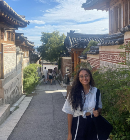 Foto de uma mulher negra, de cabelos cacheados compridos e castanhos, sorrindo. Ela usa óculos, uma camisa azul clara com nó na barriga, e uma bolsa azul escura. Ela está em uma rua estreita de inclinação leve, com casas dos dois lados, com telhados simples e detalhes vermelhos nas paredes. Algumas casas possuem arbustos na fachada. Ao fundo, um grupo de pessoas desce a rua. O céu ao fundo é azul com nuvens.